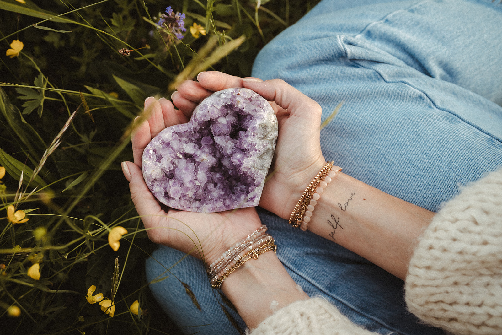 Hände halten ein Amethyst-Herz mit Heilstein-Armbändern